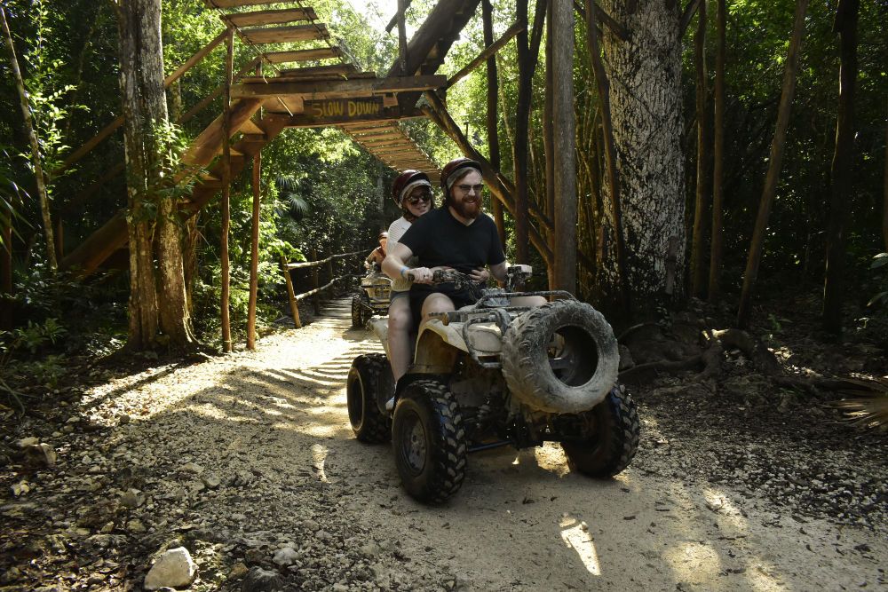 A couple is seated on an ATV, smiling for a picture. They are wearing helmets and appear to be on a jungle path, surrounded by lush greenery and trees.