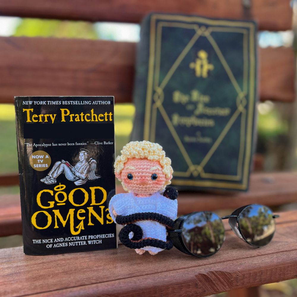 A picture that shows, from left to right, the book Good Omens (black cover with Aziraphale. The title and Terry Pratchett are written in yellow), a little crocheted Aziraphale (blond curly hair, blue eyes, white skin, wearing a white robe and no shoes) that has Crowley in his snake (black with red belly) form wrapped around him and sunglasses. Blurred in the background, there’s a green purse that looks like “The Nice and Accurate Prophecies of Ages Nutter, Witch” used in the Good Omens tv series. All the itens are on a bench. It’s possible to see that the bench has greenery around it.