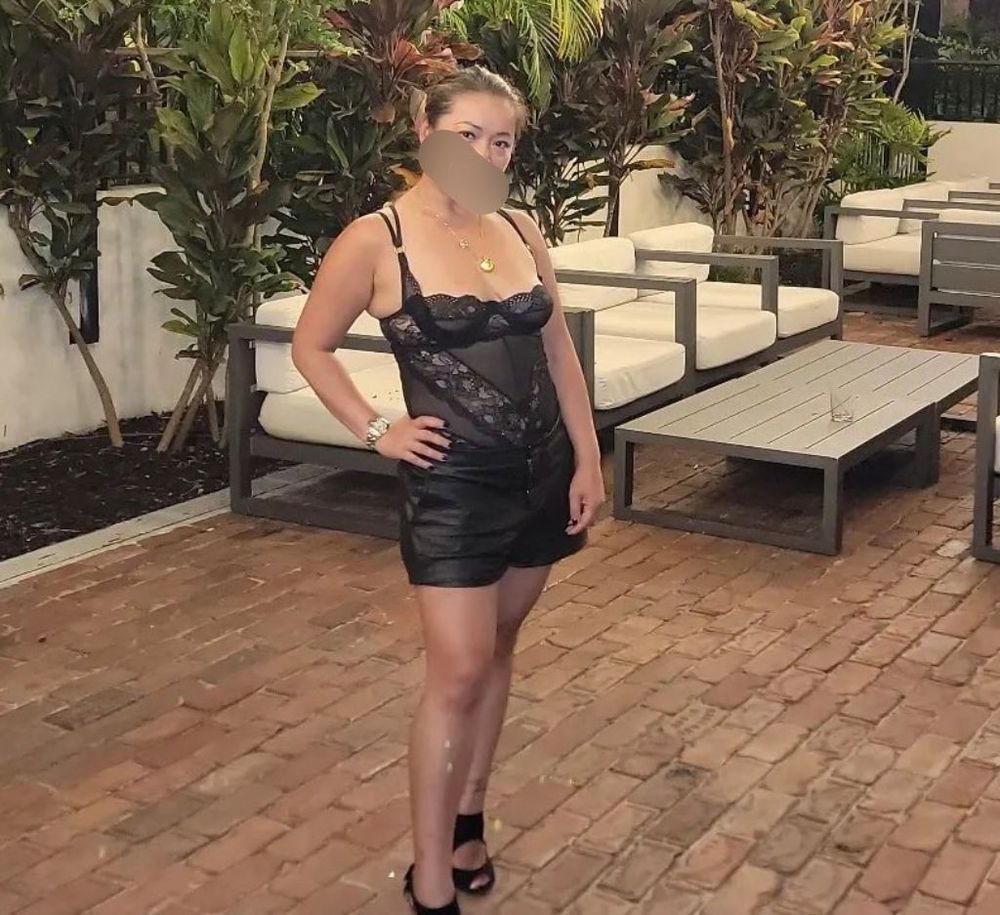 Love DeeItYi wearing a lace bodysuit with leather shorts with her right hand on her hips.  She has a seductive look.