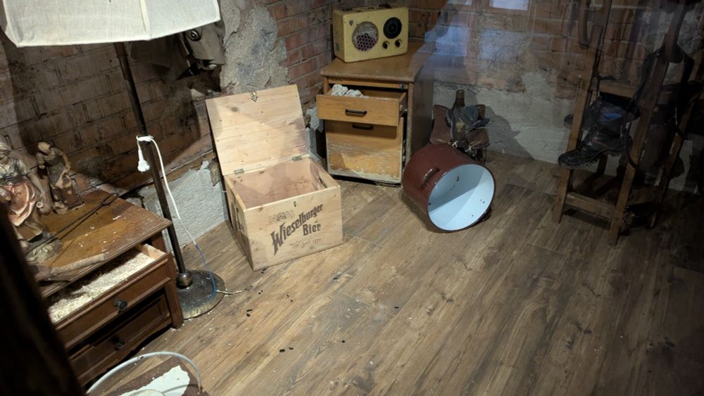 Rattenzimmer ohne Ratten, die haben alle geschlafen. In dem Zimmer sind zwei Nachtkästchen mit Schüben, eine Stehlampe, eine geöffnete Kiste, ein großer brauner Topf in Seitenlage, ein Schlitten und Schuhe zu sehen.