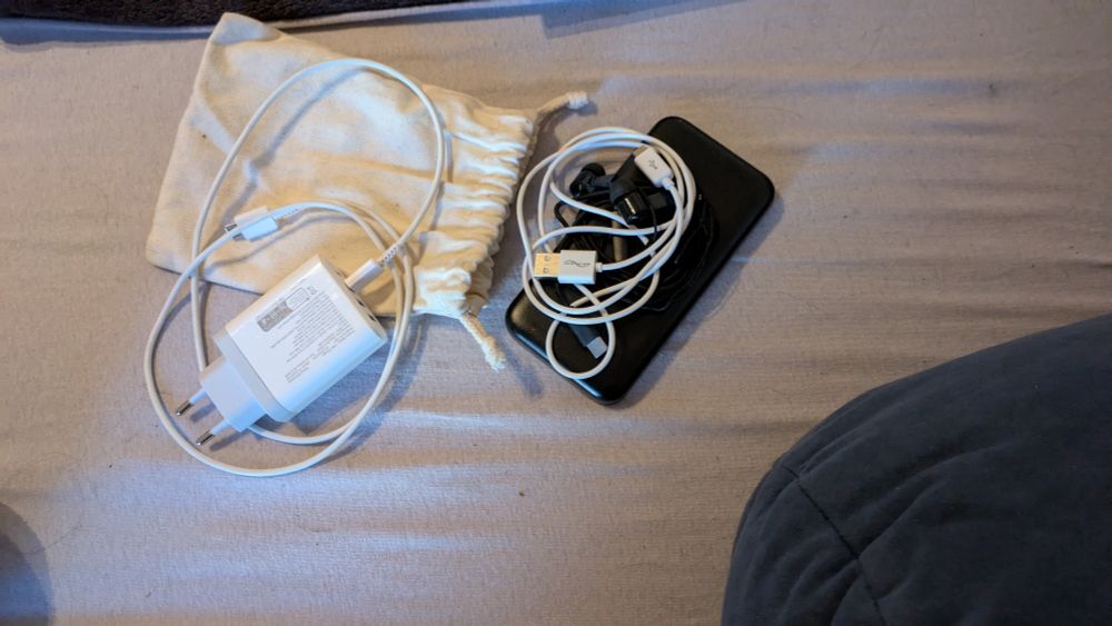 Weißes Ladekabel, helles Stoffsäckchen, eine schwarze Powerbank und weiße Kabel in Nahaufnahme auf dem Bett