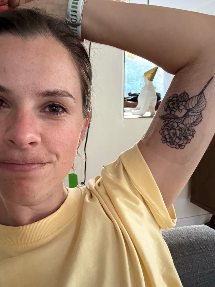 Meredith wearing a yellow shirt and showing off a brand new hydrangea tattoo on her bicep, covered by tegaderm