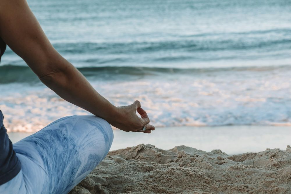  A shot of someone's arm and leg, short sleeved and wearing jeans, meditating on the sand overlooking a calm blue sea at the beach. 