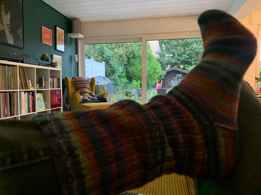 Geringelte Wollsocke im Vordergrund. Im Hintergrund ein Garten durch die Terrassentür zu sehen. 