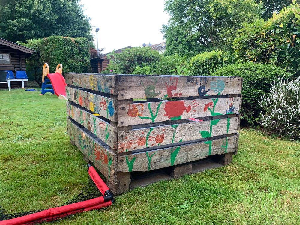 Eine grosse mit Kinderhänden bemalte Holzkiste steht auf einem Rasen. Im Hintergrund ein Gartenhaus, eine kleine Smobyrutsche und eine blaue Bank. An der rechten Seite einige Pflanzen. 