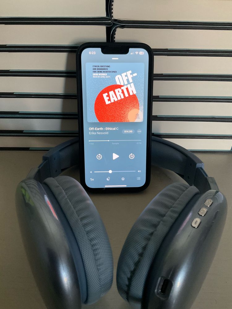 A photo of an iPhone, displaying the audiobook version of Off-Earth, leaning against a stack of books with only their edge showing. A pair of blue over-ear headphones sits beneath the phone.