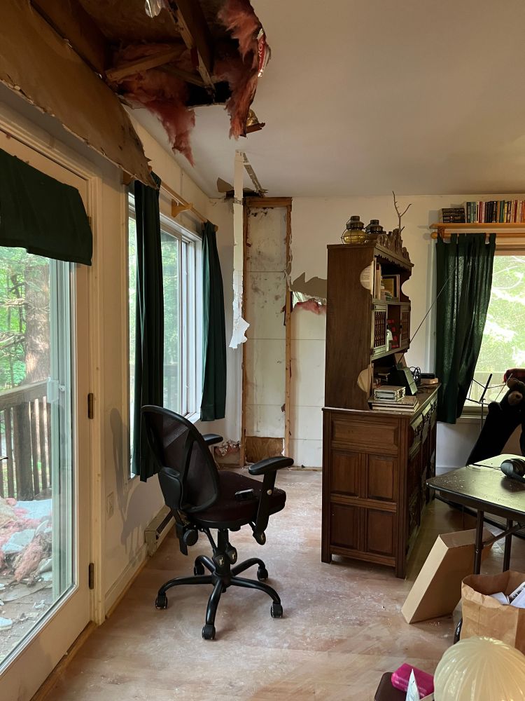 A different angle of the previous photo, showing the wall of the living room and the scattered furniture. Besides the hole in the ceiling, a large section of the drywall on one wall has been torn out and the insulation removed. A pile of pink insulation is visible through the windows, sitting outside on the porch. 