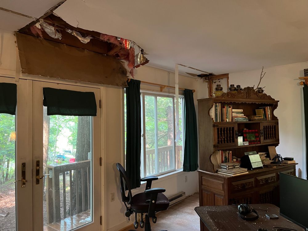 A photo of one wall of a living room. Trees and a wood porch are visible through the windows. A large chunk of the drywall in the ceiling is missing in a jagged patch, the insulation torn out. The wood floor and hastily moved table and bookshelf are covered in drywall dust. 