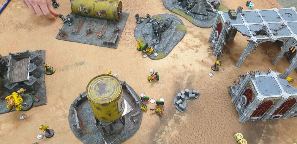 A photo of a tabletop game played with brightly-coloured yellow miniatures, and also some slightly darker ones. Some little ruined buildings, big fuel tanks/silos and other rubble scattered about a sort of ashen/desert scene.