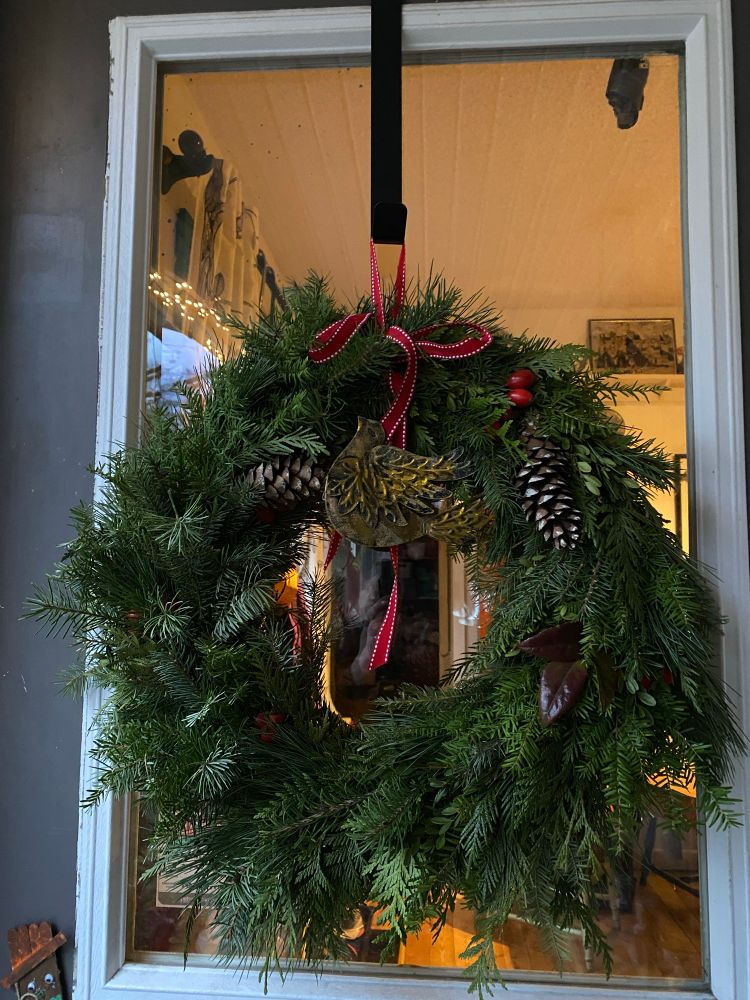 A fluffy Christmas wreath with pine cones, a red ribbon, and a gold metal bird ornament lies against a front door window, inside is warmly lit. 