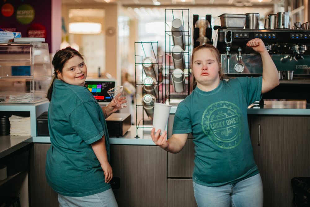 Two new coffee shop employees with cups in hand and smiles ready to make a drink in green shirts
