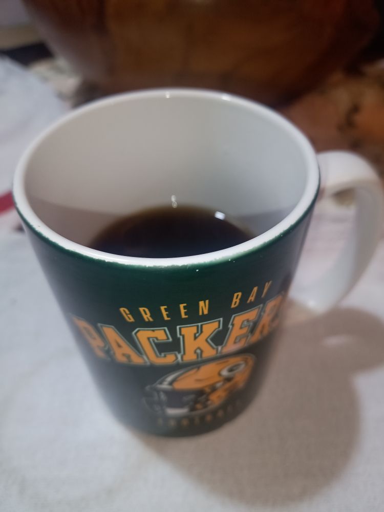 Xícara grande de cor verde por fora com o nome do time Green Bay Packers em amarelo. Interior da xícara é branco. Xícara está sob a mesa com uma toalha branca. 