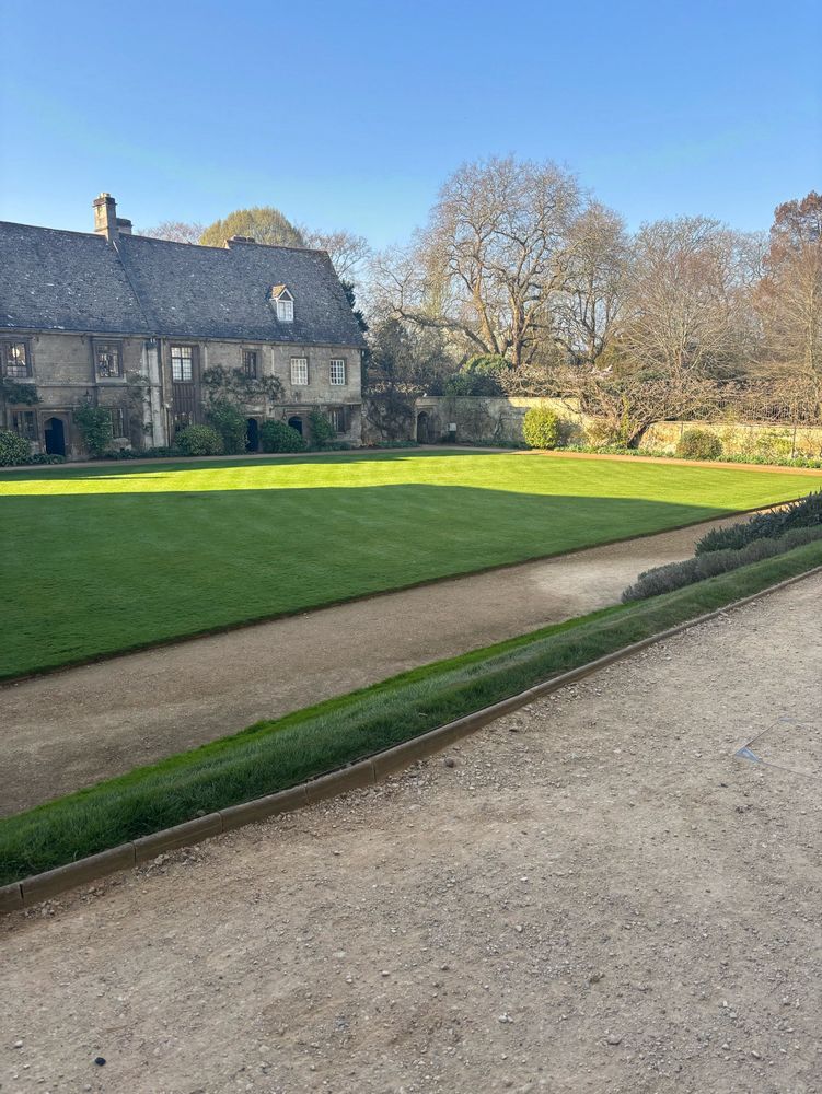 A very old college with low stone buildings with a sunny manicured lawn 