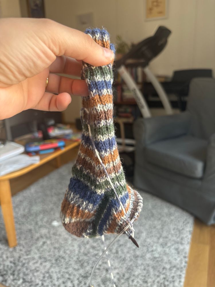 Sock with stripey pattern in blue green and brown on some circular needles. Held up by my hand- the cuff and heel turn is done but the foot is only 25% done. 
