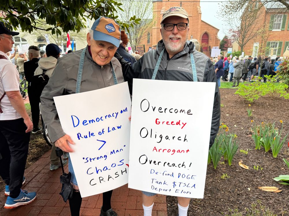Two white haired white men with light jackets and baseball caps outside with protest signs with crowd behind them.  One sign says Democracy and Rule of Law > “strong man chaos crash”.  Other says “ Overcome greedy Oligarch arrogant overreach. De-fund DoGE Tank $TSLA Deport Elon”. 