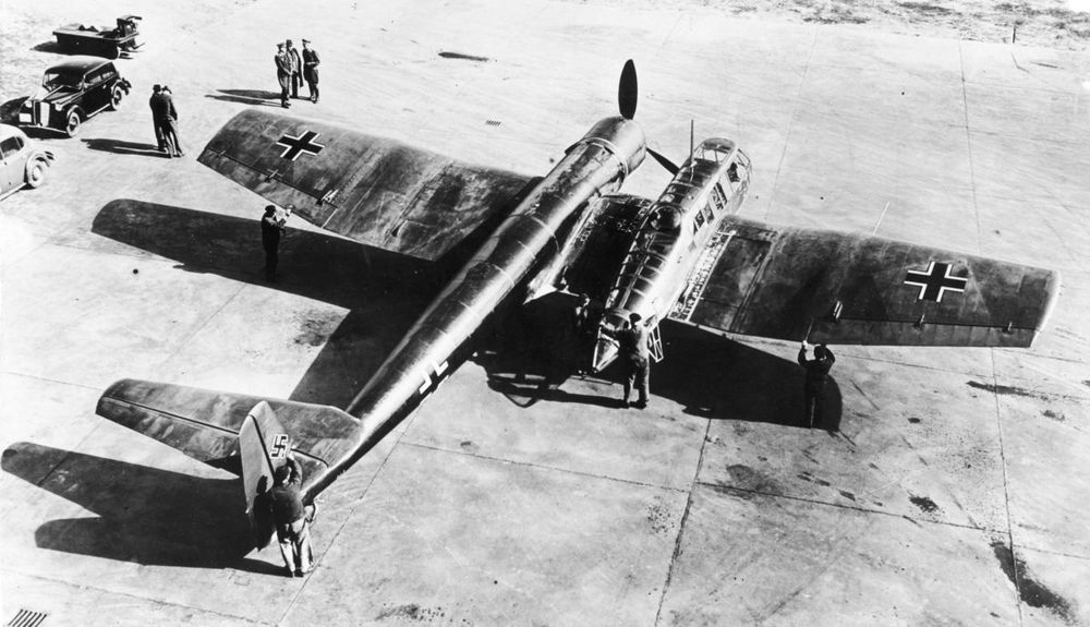 a black and white image of the Blohm & Voss BV 141. an airplane with an asymmetrical design, where the engine is on the left side of the wing and the cockpit on the right. On the tail is a swastika, used during the second world war by the German Nazis. There are handful of people looking and a few working on the plane. 