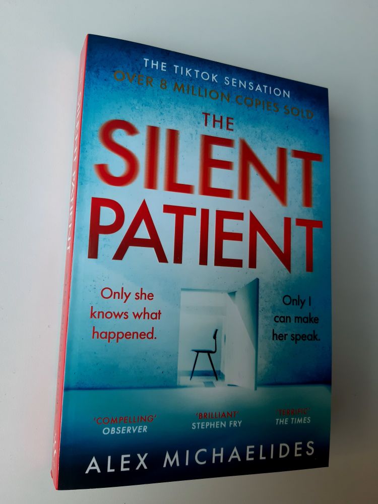 The cover of The Silent Patient by Alex Michaelides. At the top in large red text sits the title of the book. The word "Silent" is a bit blurry. In the middle of the cover is a photo of an open door with in the room behind it stands a simple chair. At the left side reads the text "Only she knows what happened." in red. At the right side reads the text "Only I can make her speak." in black. At the bottom sits the author's name. There are multiple text of praises from different people and media outlets spread on the cover. 