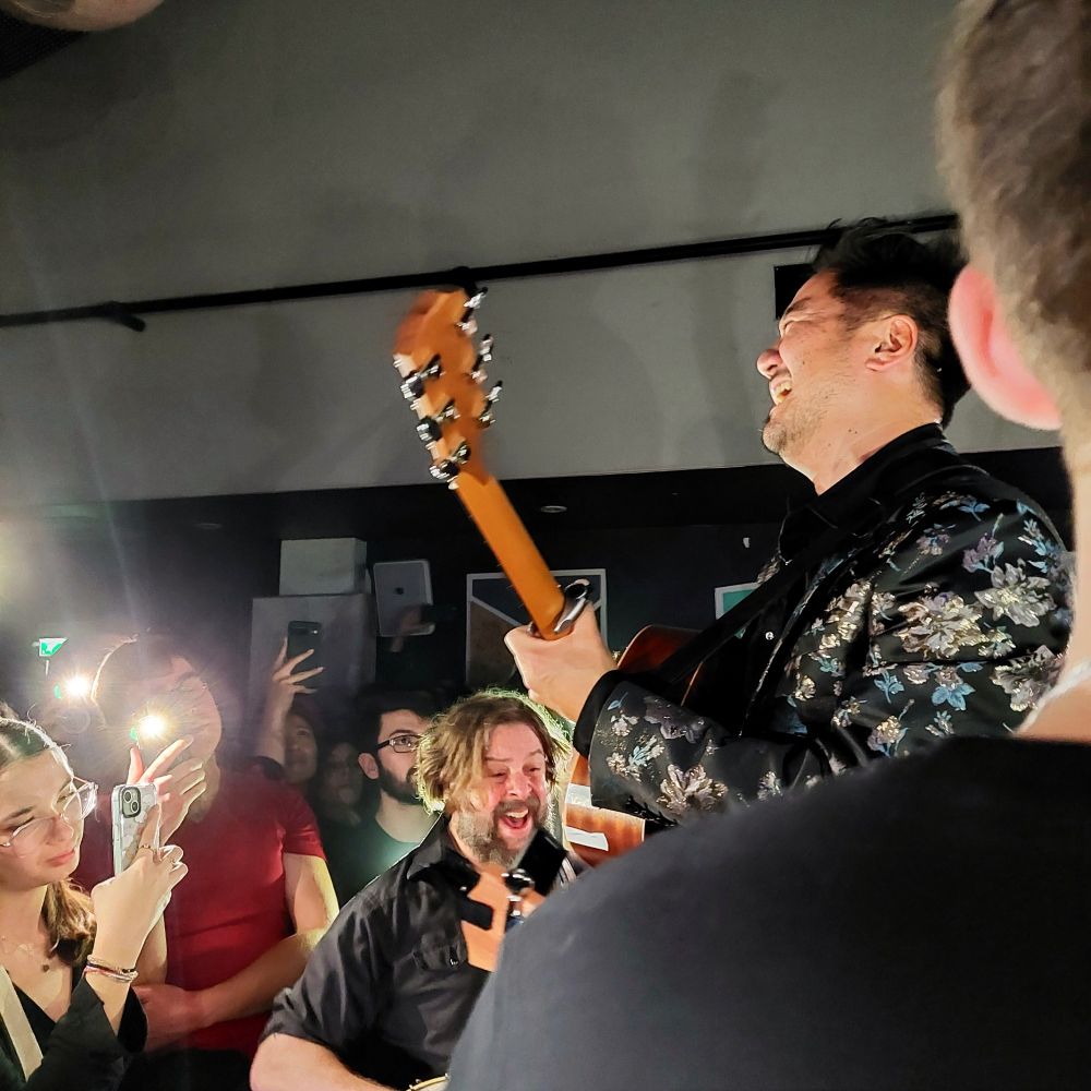Kishi Bashi playing an acoustic guitar in the middle of the audience while standing on a chair with Tall Tall Trees sings harmonies below him. 

People have their phone lights on giving a bright contrasting lighting to the scene.