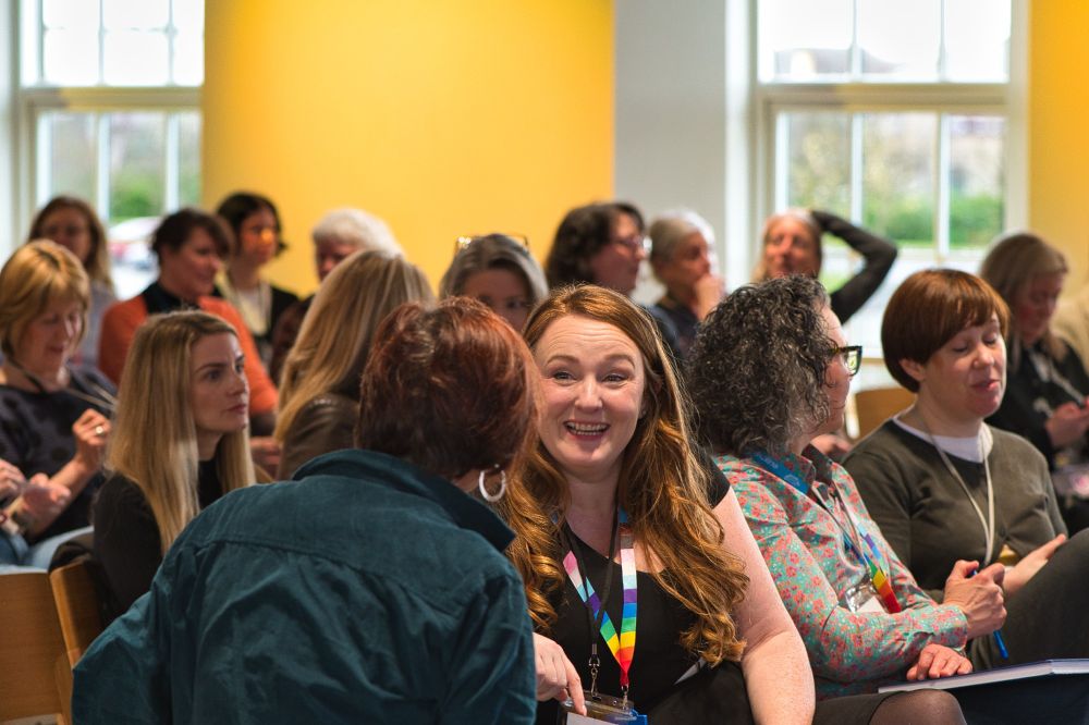 group of people in a conference setting in rows of chairs. People are talking to each other and looking ahead. In the foreground, two people are talking and smiling.