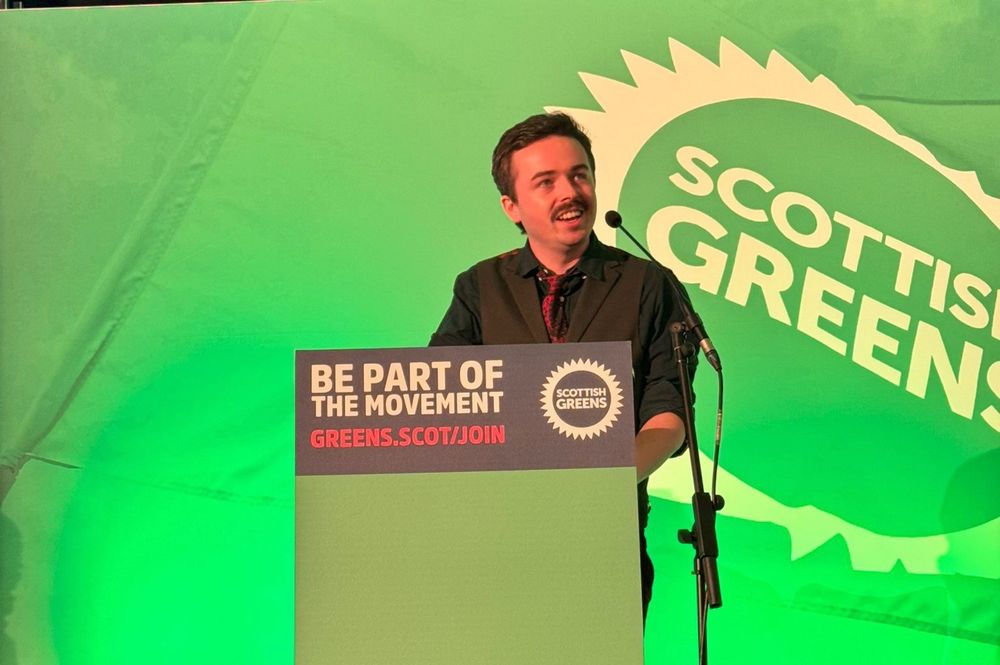 A smiling Anthony behind a stand and infront of the Scottish Greens logo