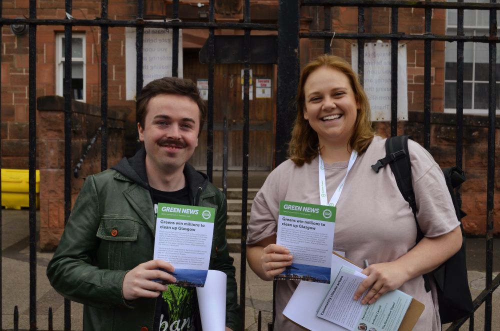 Anthony & Kate smiling to camera holding leaflets, school is behind them 