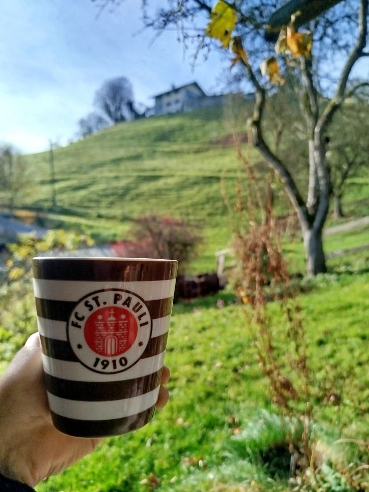Foto: FC St. Pauli Kaffeetasse, im Hintergrund Hügellandschaft mit Baum und Haus