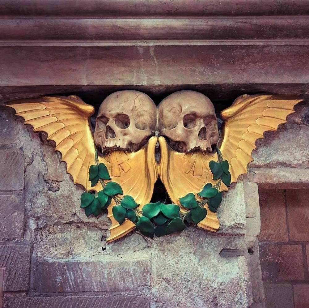 Two carved pale stone skulls, looking away from each other, framed by golden painted wings and a garland of green painted leaves