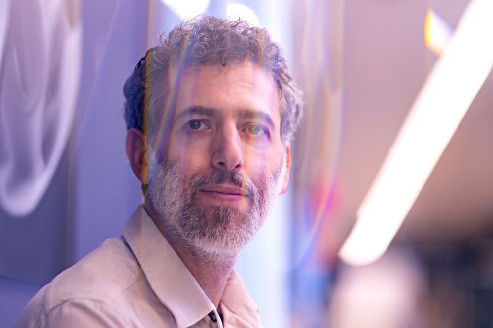 A man with a salt/pepper beard looking at the camera. the lights reflecting on the lens of the camera looks like his face is in a bubble. 