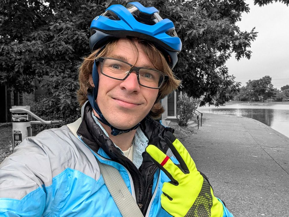 A selfie of Robin in her colorful high vis biking gear throwing a peace sign, with the trees and pond at Petaluma's Lucchesi Park in the background in black and white.