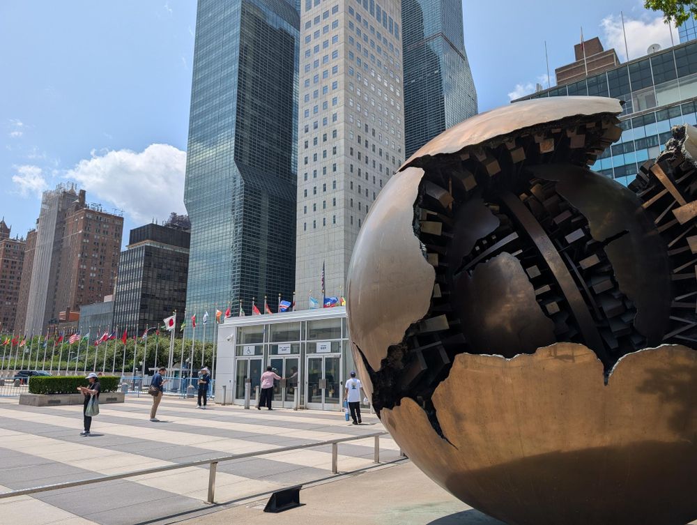 A photo from a plaza inside the United Nations Headquarters, with the iconic line of flags set against a backdrop of towers on a sunny summer day in New York. In the foreground on the right is one of the bronze sculptures designed by the artist.