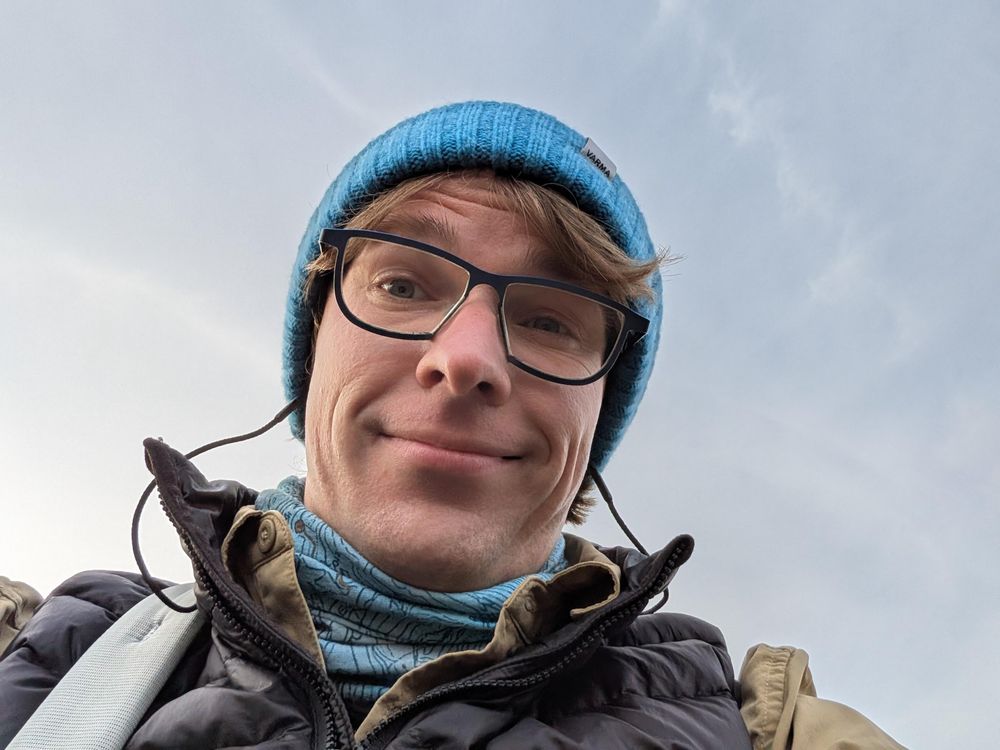 A selfie of Robin layered up on a chilly late spring evening, looking down into the camera with the sky filling the background. She's rocking a bright blue knit beanie, blue and brown gaiter, and a black vest over a brown shirt. 