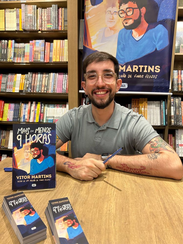 Vitor Martins posando com seu livro Mais ou Menos 9 Horas em exposição e alguns marcadores de livro em cima da mesa