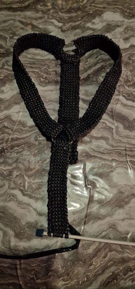 Image shows a V Neck harness, knitted in a black shiny pleather material, with metal rings in the front and back. Resting on a marble vinyl covered table.