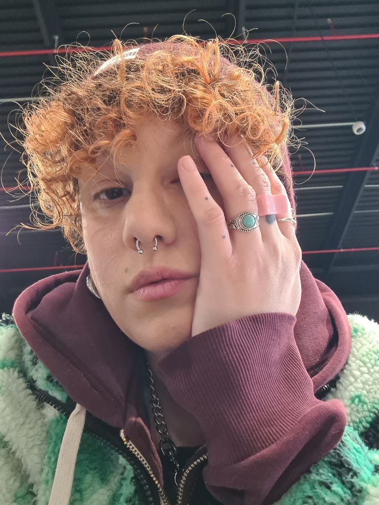 Una persona abrigada porque hoy nevó! 
La mano en la cara con varios anillos. Llevo un septum. Un gorro, una sudadera con capucha de color granate y una chaqueta verde polar.
Es un selfie.