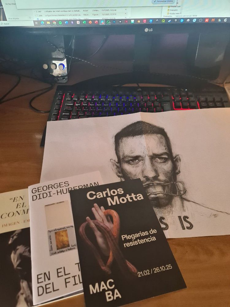 Fotografía de una mesa de escritorio. Sobre el teclado del ordenador se ve un libreto desplegado con una ilustración de Carlos Motta donde aparece el artista retratado. No se lee, pero debajo de su rostro pone AIDS IS. También un folleto de su exposición. También hay otros dos libretos de la expo de Didi-Huberman. 