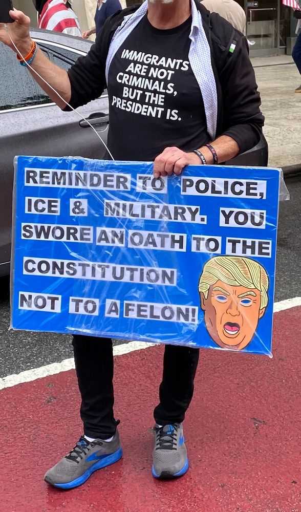 Man wearing a t-shirt that says: "Immigrants are not criminals, but the president is." He's holding a sign that says: "Reminder to police, ICE, and military. You swore an oath to the Constitution, not to a felon!"