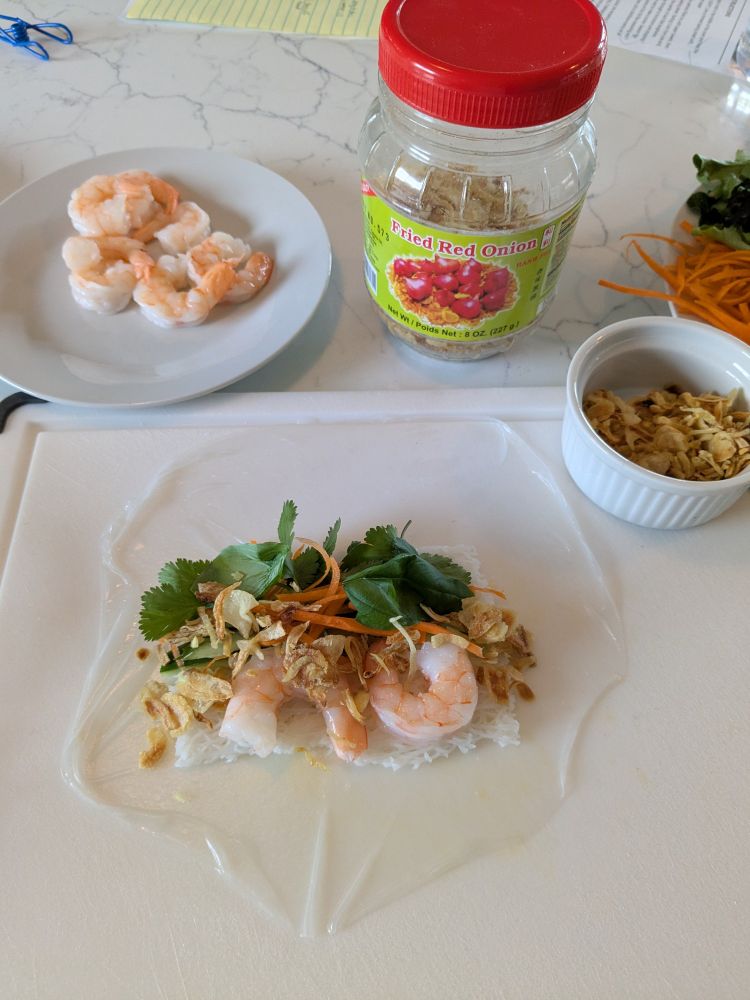 Salad roll wrapper with herbs, shrimp, fried shallots on cutting board.
