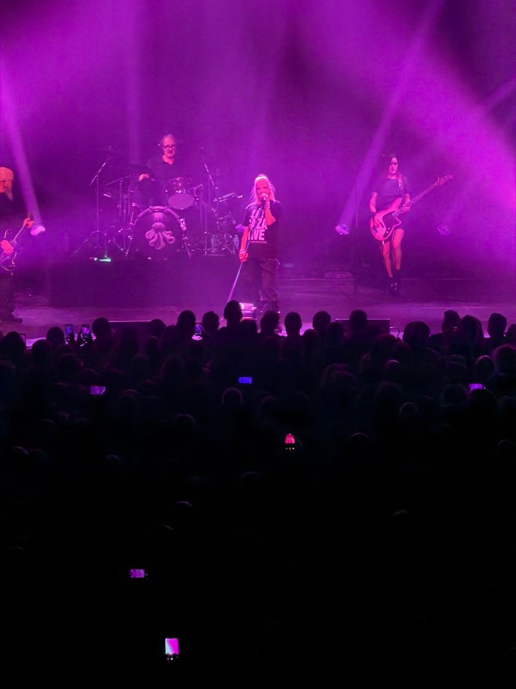 The image shows a live concert performance on a stage bathed in bright purple lighting. A rock band with five members is performing: a lead singer stands at the center holding a microphone stand, a drummer is seated behind a drum kit with an octopus logo, a guitarist is on the left, and a bassist is on the right. The audience is in the foreground, mostly in shadow, with several people holding up glowing smartphones to capture the performance. The beams of stage lights cut through the purple haze, creating a dramatic atmosphere.