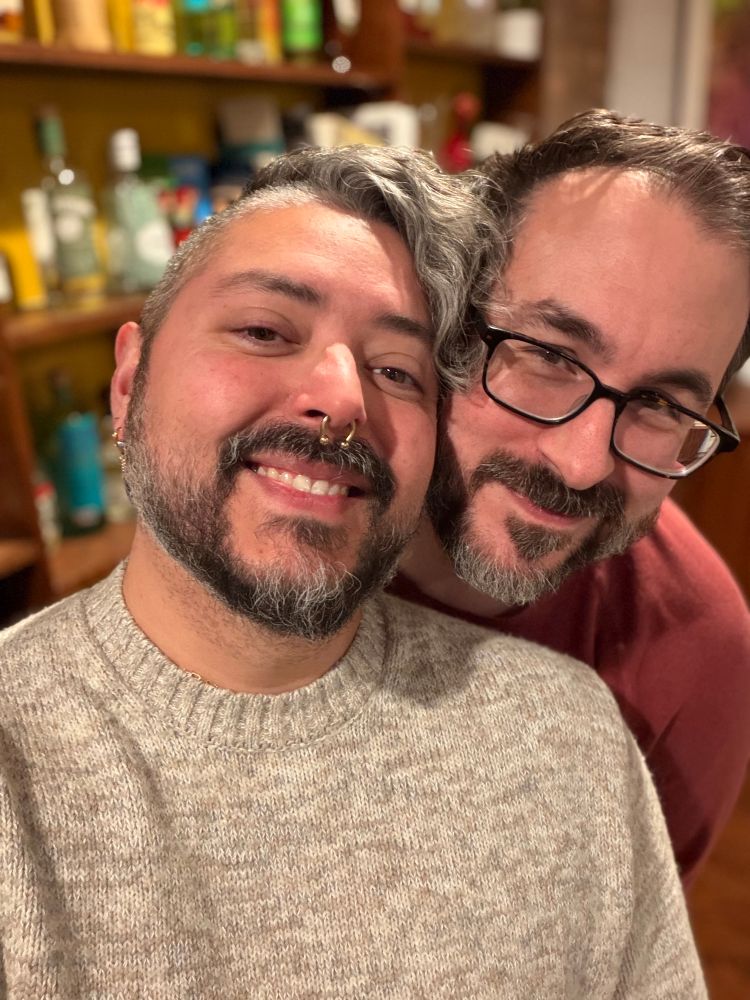 Two smiling people are posing for a close-up selfie indoors. The person in front has short gray hair with a wavy section falling to one side, a beard, and a septum piercing. They are wearing a light gray knit sweater. The person behind them has short dark hair, a beard, and is wearing glasses and a red sweater. They are leaning their head close to the person in front. The background shows shelves with bottles and various items, slightly out of focus.