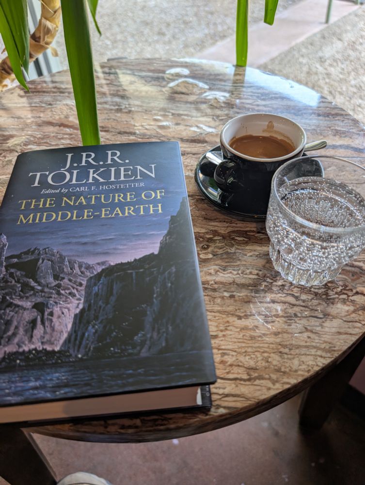 Close up of a copy of "The Nature of Middle-earth" by J.R.R. Tolkien next to a cup of espresso and sparkling water on an end table.