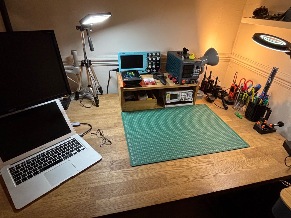 Clean desk. Computer on the left, scope and other test gear plus obligatory green mat in the mid, various VHF handsets and pens and the like on the right.