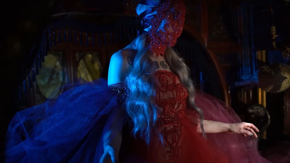 A still of Lingua Ignota from her music video for "Perpetual Flame of Centralia". She's wearing an ornate red gown with excessive beadwork and diaphanous layers, with a matching full-face mask. Her chest tattoo is visible, framed by the top of her dress and the bottom of her mask.