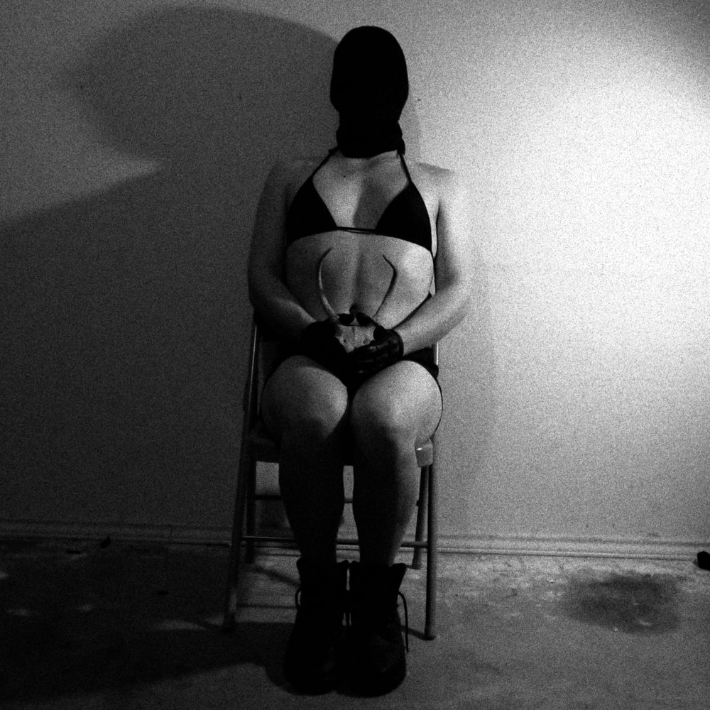 A black and white photograph of Ash sitting on a folding steel chair against a featureless wall in a dingy garage. She is wearing a black bikini, ragged black leather boots, black leather gloves and a black bag over her head. Though her body looks soft, her breasts are still small. She is sitting demurely, with her legs together, back straight. She is holding the skull of a young male deer in her lap. The horns of the buck frame her solar plexus perfectly. There is a single light source on the right, with the light breaking suggestively over her body and casting a broad silhouette across the wall to her left.