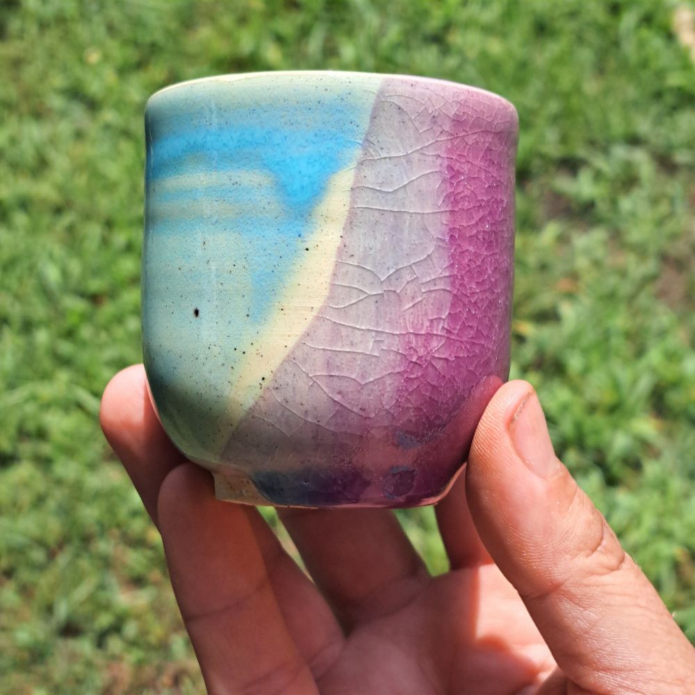 A small handleless cup made of white stoneware clay. It is glazed in robin egg blue and a gloriously crazed purple