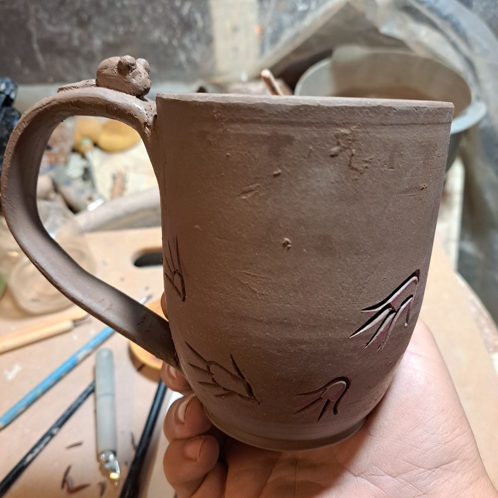 A clay mug at leather hard stage.  There are stylized rat tracks carved on it and a little sculpture of a rat on the handle.