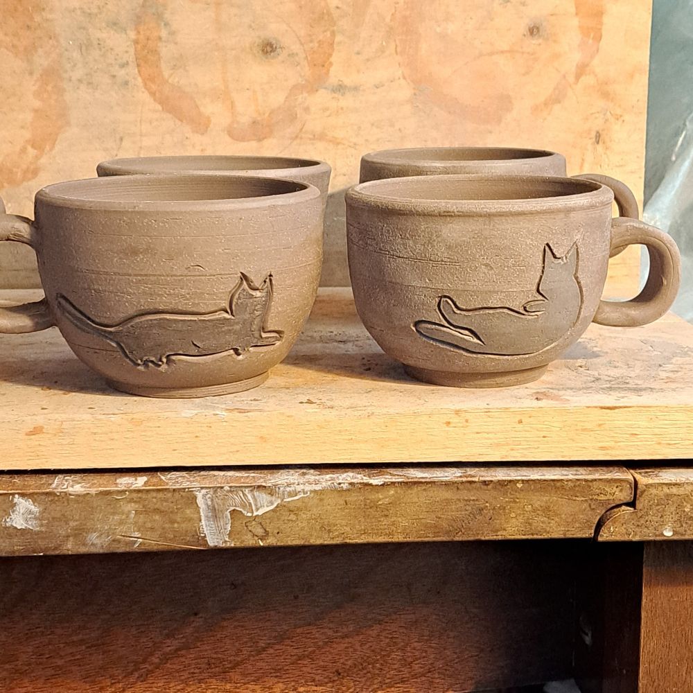 Leather-hard greenware teacups carved with sleepy cat silhouettes. 