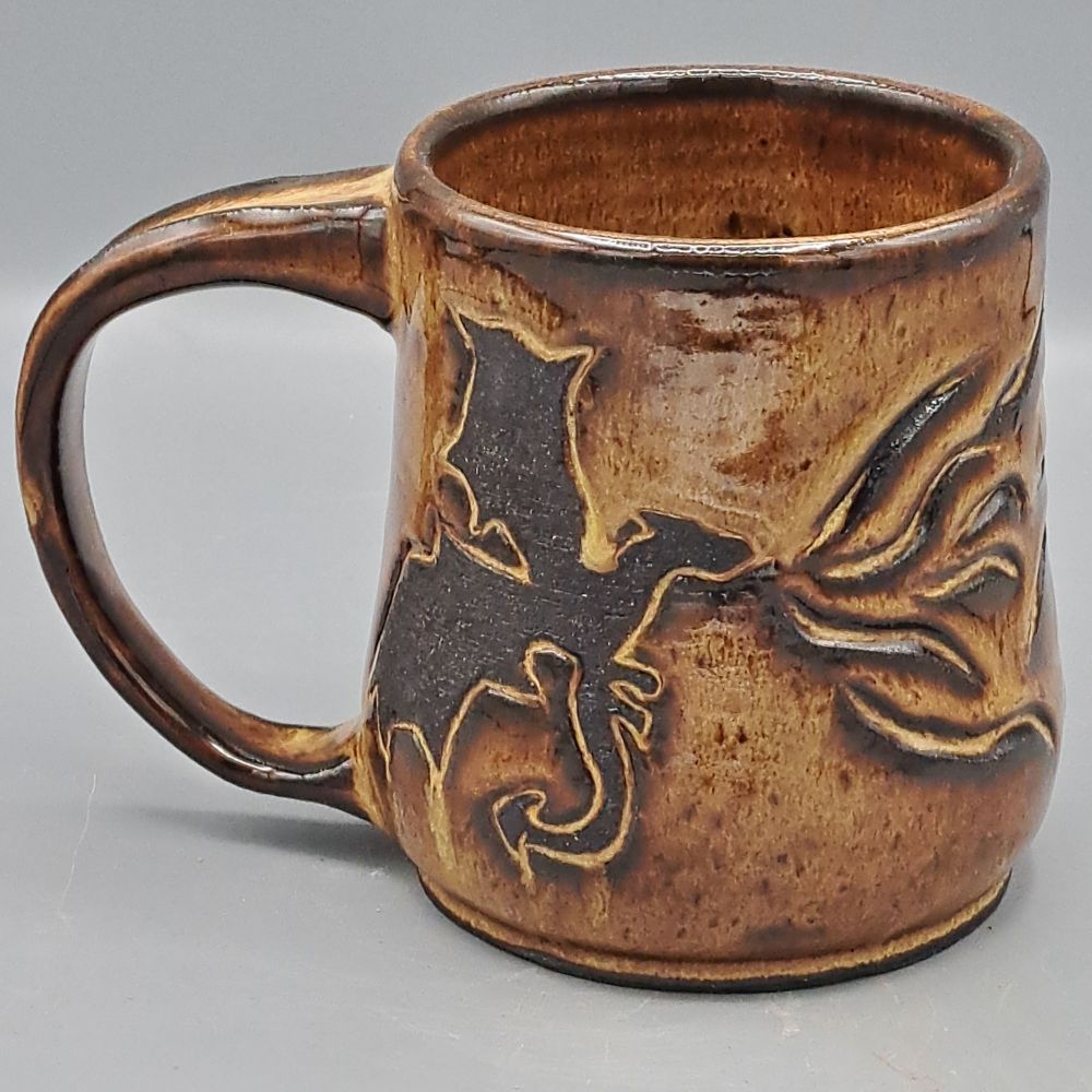 A mug made of black clay and glazed in golden/bronze. A dragon blowing flames is carved and resisted in raw black clay.