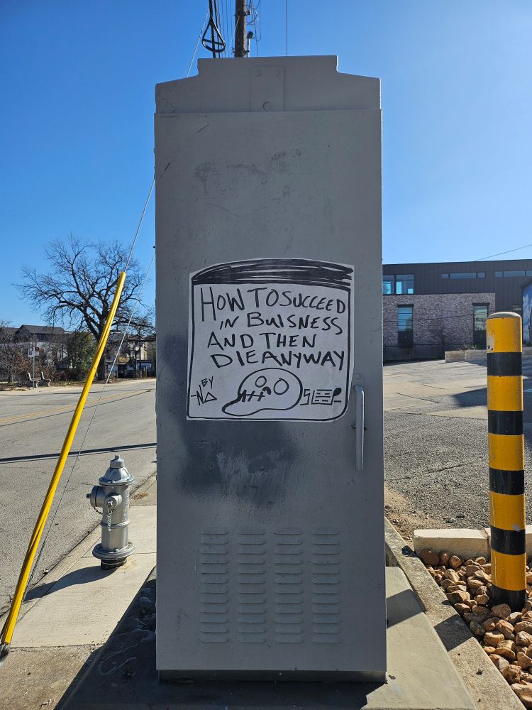 A big old piece of sticker graffiti on some kind of electrical housing on a street corner. The sticker has a rough drawing of a book in black marker. The cover features a skull, and the title is "how to succeed at buisness [sic] and then die anyway."