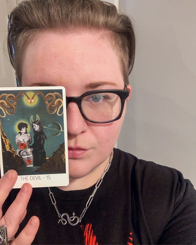 Person with glasses holds The Devil tarot card over one eye, wearing black shirt and snake necklace.