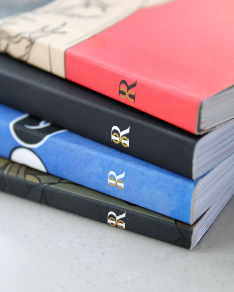 A close-up image of four stacked books with colorful spines in red, black, blue, and green. Each spine features a small, elegant gold and white symbol resembling a mirrored pair of lowercase “a” characters or a stylized infinity sign. The books appear to be high-quality print editions, resting on a light gray surface.
